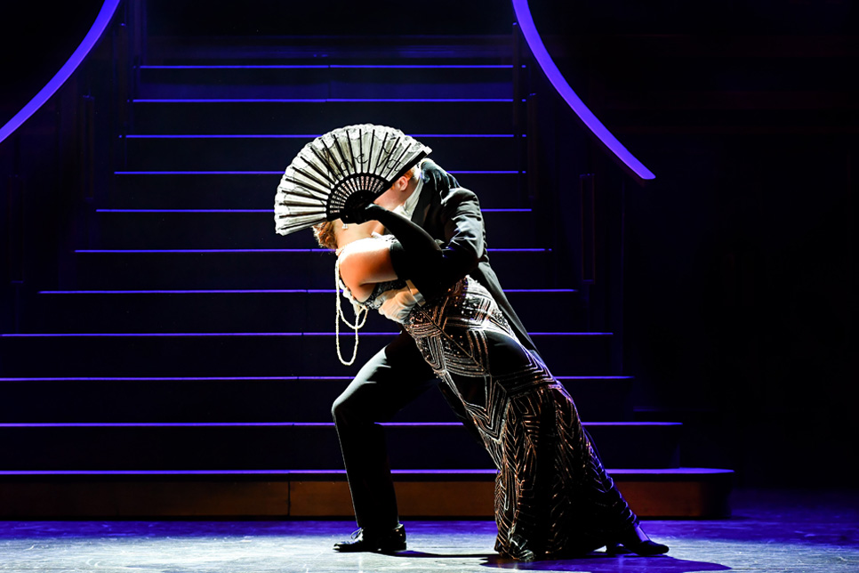 Students dressed in 1920s fashion, kissing on stage, hidden behind a fan, in front of a staircase.